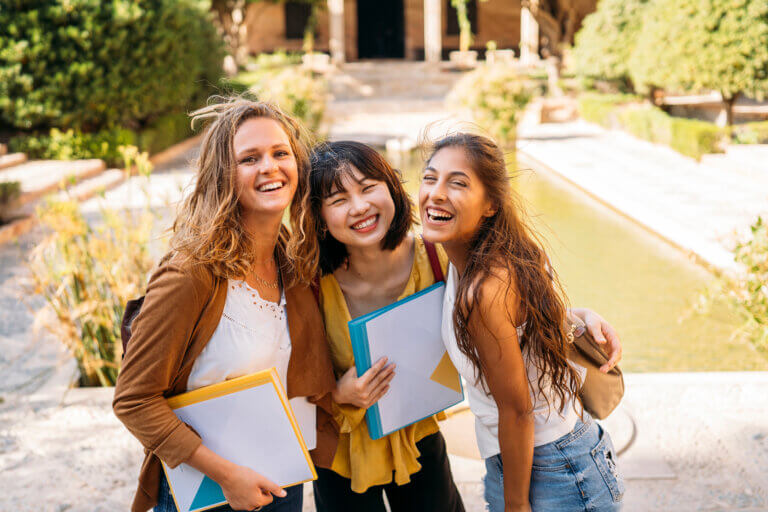 Three students standing close together outdoors beside a narrow water feature on a landscaped campus walkway. Each person is holding folders or notebooks, and all are facing the camera. Trees, shrubs, stone pathways, and a building entrance appear in the softly blurred background, illuminated by warm sunlight.