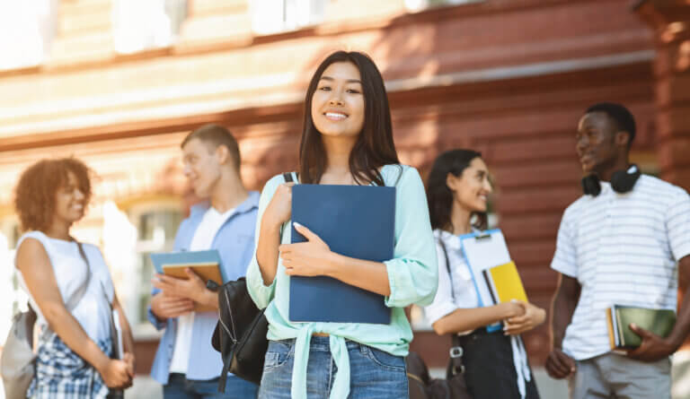A person standing outdoors in front of a campus-style building holds a blue binder and wears a backpack. Several other students carrying notebooks and folders stand nearby in the background. The scene is brightly lit by warm sunlight, with the central subject in focus and the surrounding students softly blurred.