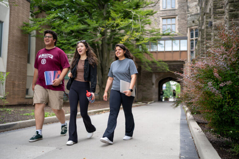 Three students walk along a tree-lined campus pathway between stone academic buildings. One carries textbooks, another holds a water bottle, and a third carries a laptop. An arched passageway is visible in the background.