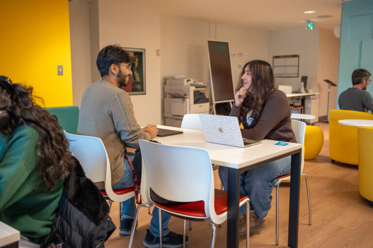 Students chat around a table in a bright shared office, with a woman using a laptop, colorful chairs, and others working in the background.