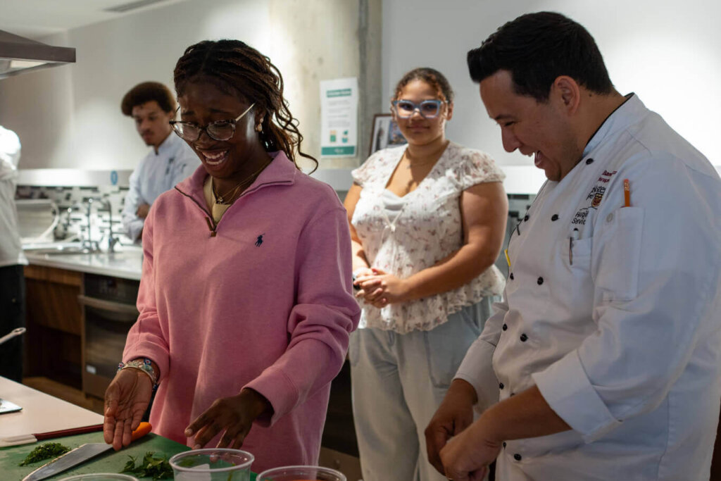 Cooking class with a diverse group preparing ingredients at a kitchen counter; a chef in white observes as participants work together.