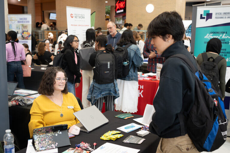 Busy career fair with diverse attendees browsing nonprofit booths; a smiling vendor in yellow speaks with a visitor carrying a backpack.