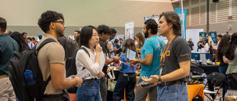 Busy career fair scene with diverse students networking, exchanging brochures, and speaking with recruiters at crowded exhibition booths.