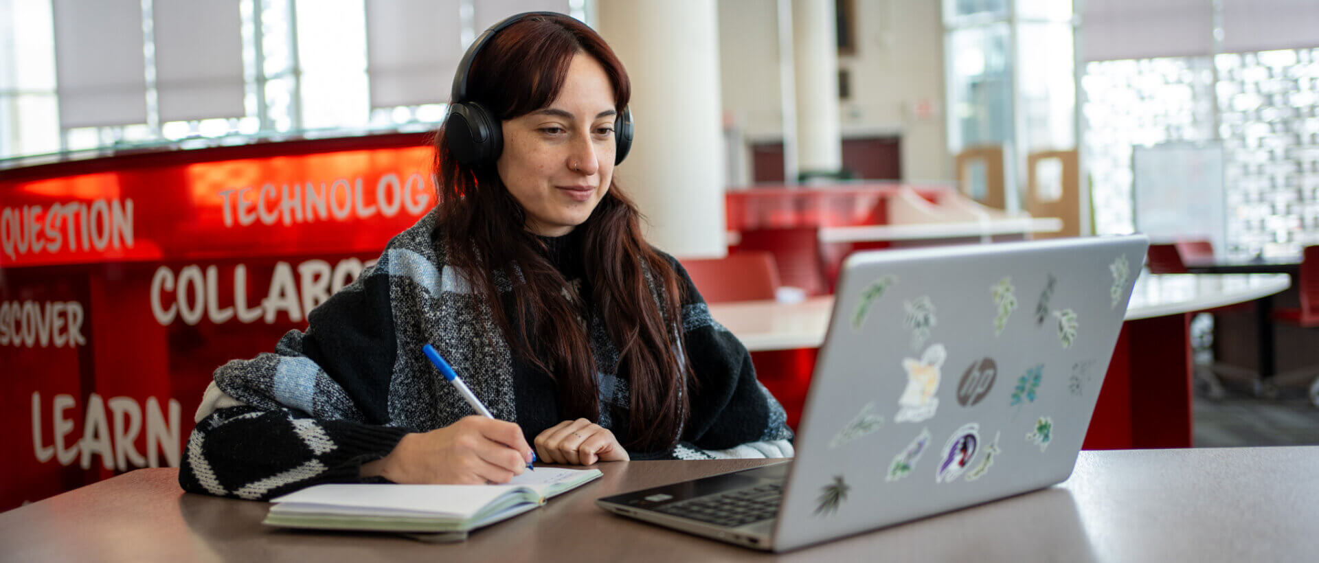 Young woman wearing headphones writes in a notebook beside a sticker-covered laptop in a bright library, creating a focused, productive mood.