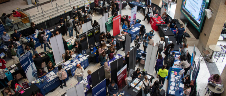 Overhead view of a busy career fair, with crowds browsing colorful company booths, banners, tables, and displays in an energetic convention hall.